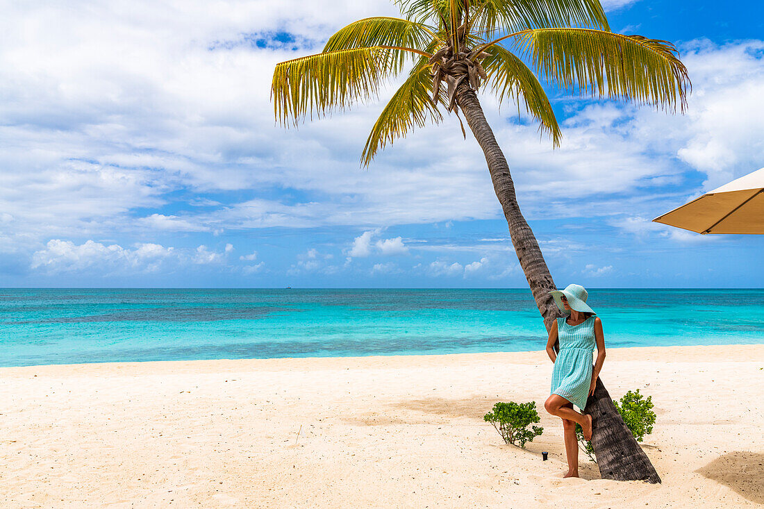 Frau in türkisfarbenem Kleid lehnt an einer Palme an einem idyllischen tropischen Strand, Barbuda, Antigua und Barbuda, Karibik, Westindische Inseln