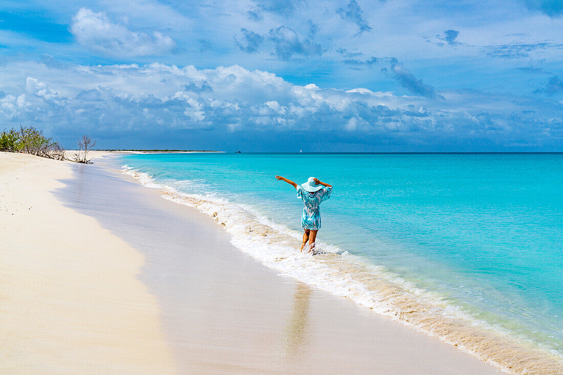 Rückansicht einer fröhlichen Frau, die den Strohhut hält, während sie mit den Wellen spielt, Barbuda, Antigua & Barbuda, Karibik, Westindische Inseln