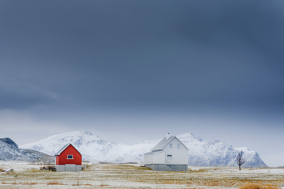 Gewitterwolken über vereinzelten Holzhäusern im Winterschnee, Flakstad, Bezirk Nordland, Lofoten Inseln, Norwegen