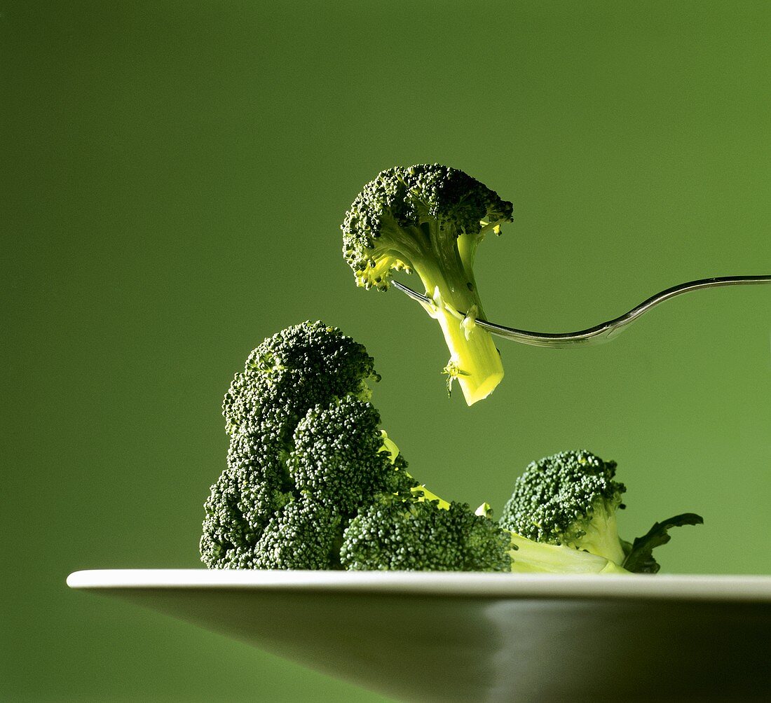 Broccoliröschen auf Gabel über Teller mit Brokkoli