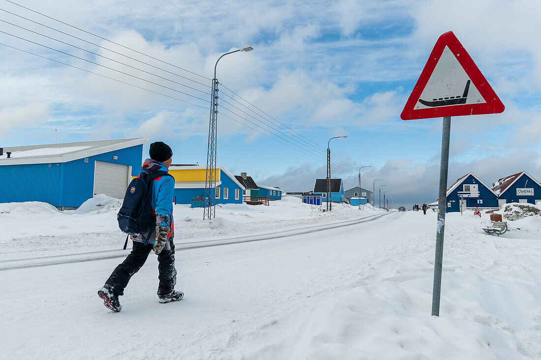 Ein Kind, das auf einer schneebedeckten Straße nach Hause geht und dabei an einem Warnschild für Hundeschlitten vorbeikommt. Ilulissat, Grönland.