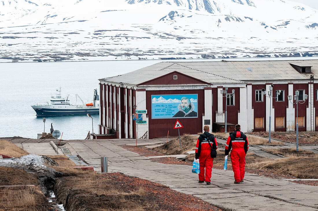 Männer in arktischer Schutzkleidung gehen auf ein verankertes Schiff zu. Barentsburg, Insel Spitzbergen, Svalbard, Norwegen.