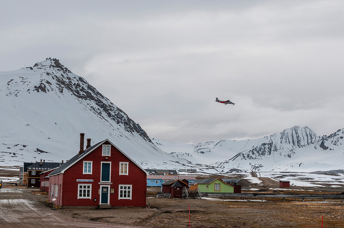 A plane flies above multicolored buildings and mountains at Ny-Alesund. Ny-Alesund, Kongsfjorden, Spitsbergen Island, Svalbard, Norway.