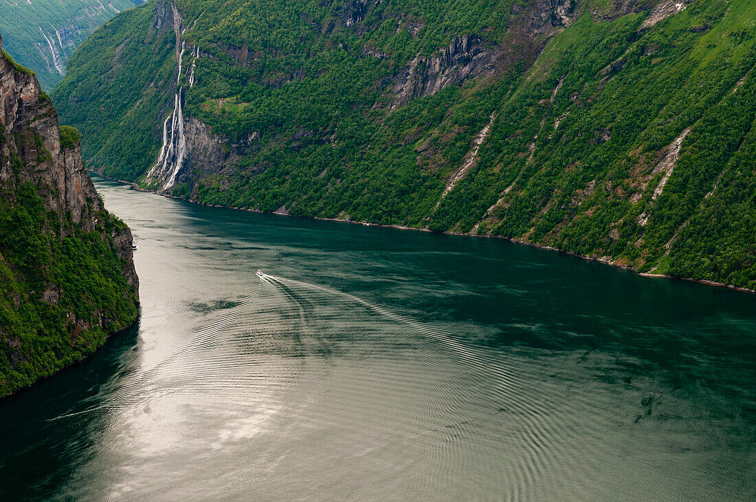 Das Kielwasser eines Bootes kräuselt den Geirangerfjord, der sich durch waldbedeckte steile Klippen und Berge windet. In der Ferne sind die Wasserfälle der Sieben Schwestern zu sehen. Geirangerfjord, Norwegen.