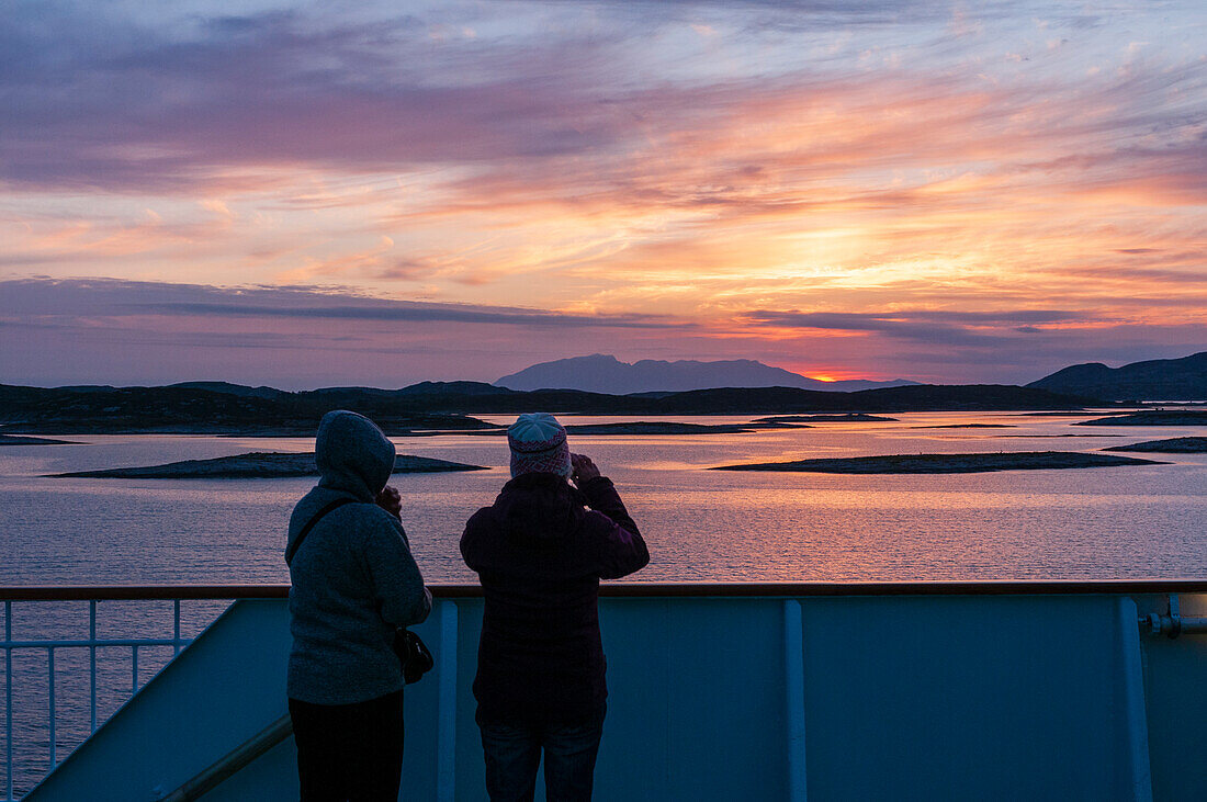 Touristen an Bord eines Kreuzfahrtschiffs betrachten den Sonnenuntergang hinter den silhouettierten Inseln in der Norwegischen See. Norwegisches Meer, Bronnoy, Nordland, Norwegen.
