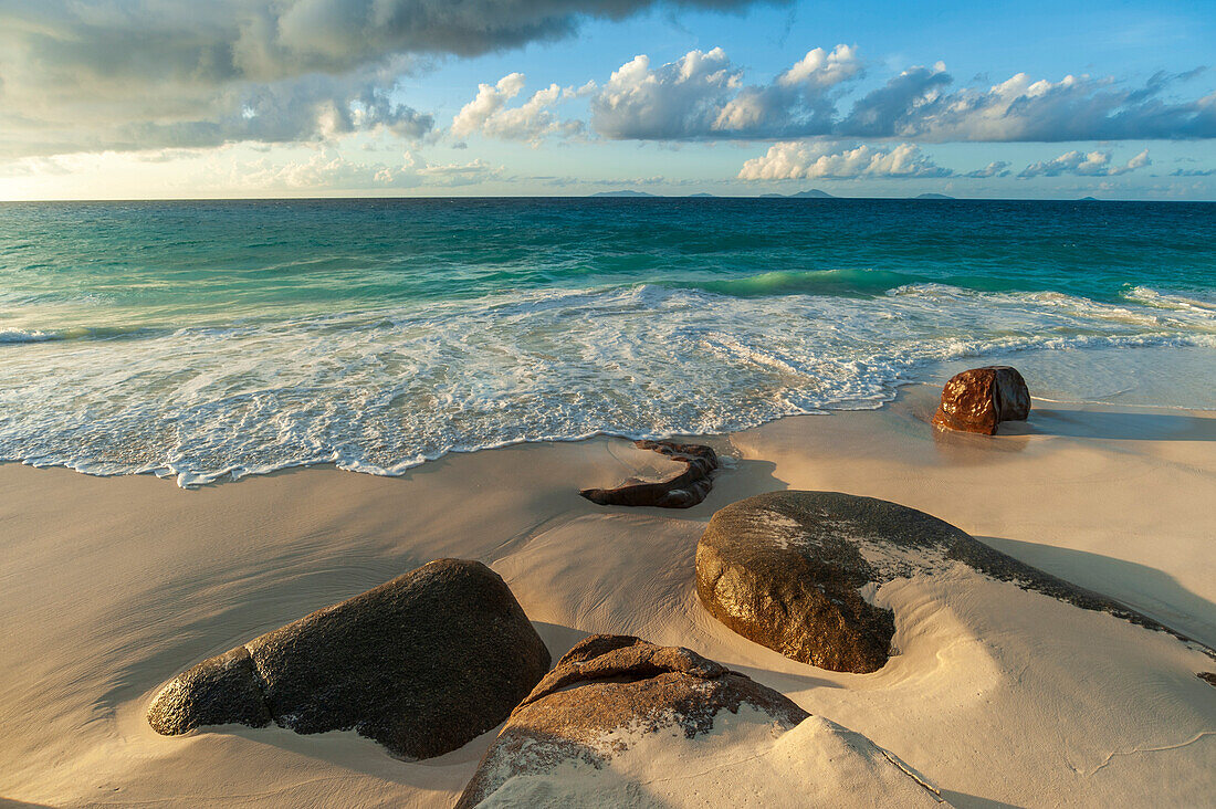 Die Brandung schlägt an einem tropischen Strand gegen Felsbrocken, die im Sand vergraben sind. Strand Anse Victorin, Insel Fregate, Republik Seychellen.