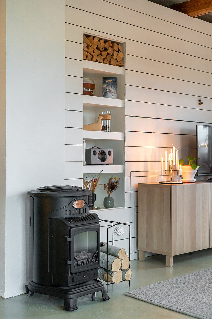 Black stove next to TV cabinet in front of white wood panelling with integrated shelf