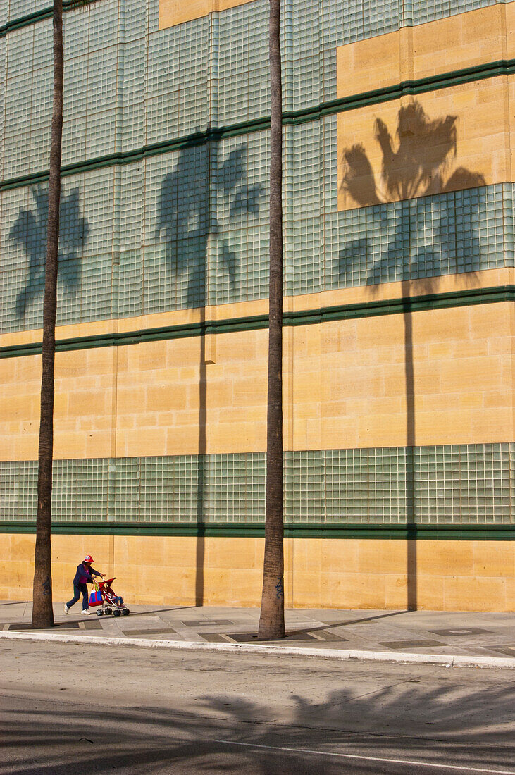 Schatten und Stämme von Palmen vor dem Los Angeles County Museum of Art, mit einer Frau, die einen Kinderwagen auf dem Bürgersteig schiebt, Wilshire Blvd, Los Angeles, Kalifornien.