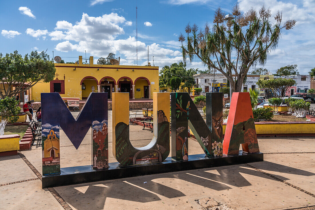 The colorful town sign in the plaza for Muna, Yucatan, Mexico. In the background is the yellow municipal palace or city hall.