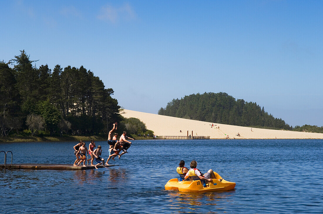 Ein Paar in einem Paddelboot und eine Gruppe Jugendlicher, die auf dem Cleawox Lake im Honeyman State Park an der Küste von Oregon vom Boot springen.