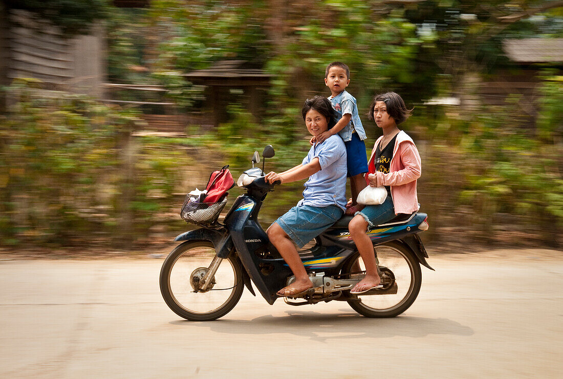 Family riding motorcycle through village along the Mae Taeng River in rural Chiang Mai Province, Thailand.