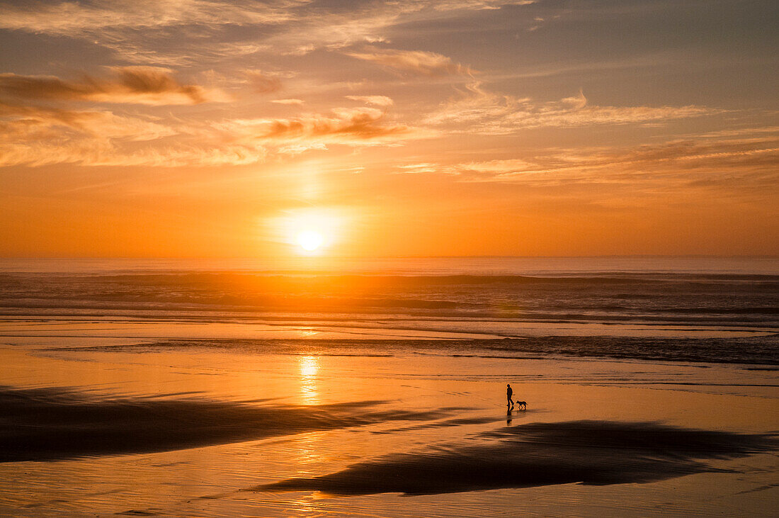 Frau geht bei Sonnenuntergang mit ihrem Hund am Tillicum Beach an der Küste von Oregon spazieren.