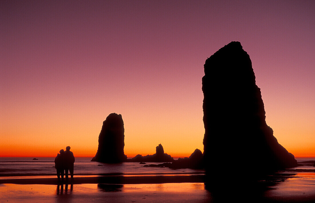 Haystack Rock & Sea Stacks bei Sonnenuntergang mit Paar am Strand; Cannon Beach, Küste von Oregon.