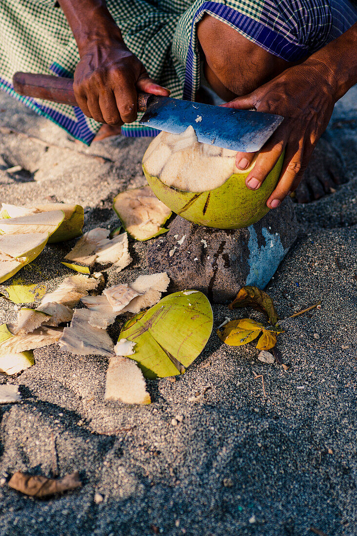 Nahaufnahme eines Mannes, der eine Kokosnuss am Nippah Beach auf der tropischen Insel Lombok, Indonesien, öffnet. Dieses Bild eines Mannes, der eine Kokosnuss auf der tropischen Insel Lombok öffnet, wurde am Nippah Beach im Westen der Insel aufgenommen. Nippah Beach ist ein atemberaubender, tropischer Strand mit wunderschönem weißen Sand an der Westküste von Lombok. Das tropische Inselparadies Lombok in der Region West Nusa Tenggara in Indonesien beherbergt einige der besten Strände der Welt, und der Nippah Beach ist nur ein Beispiel dafür.