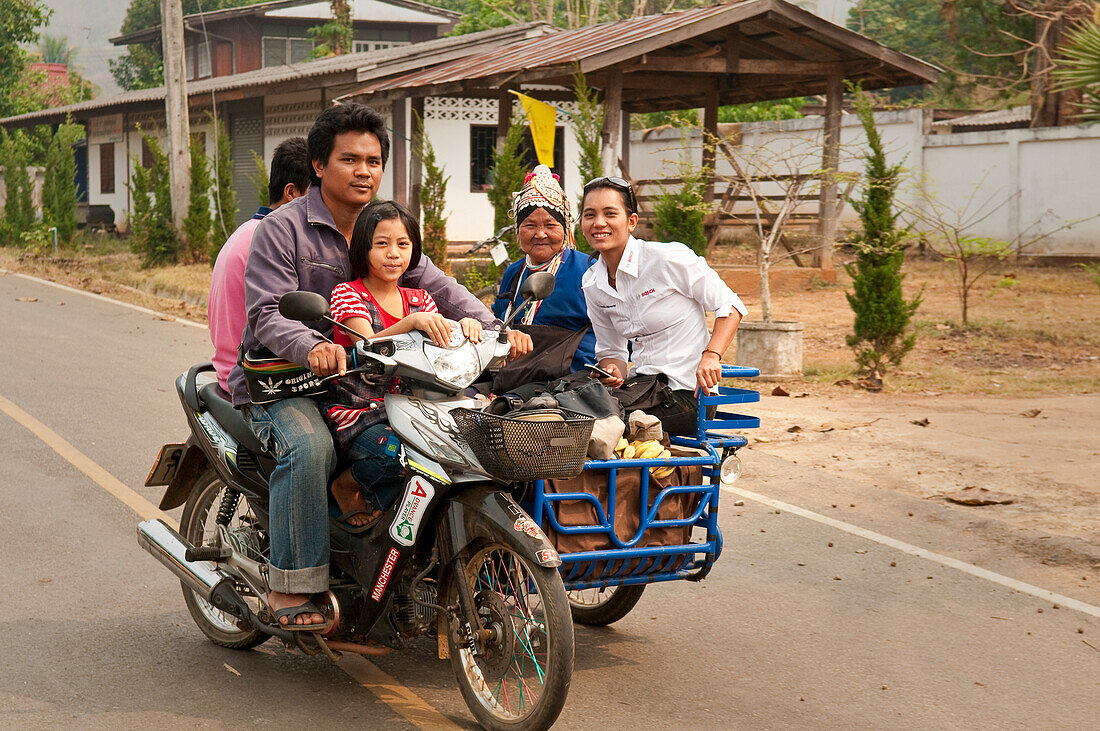 Family riding motorcycle and sidecar through village along the Mae Tang River in rural Chiang Mai Province, Thailand.