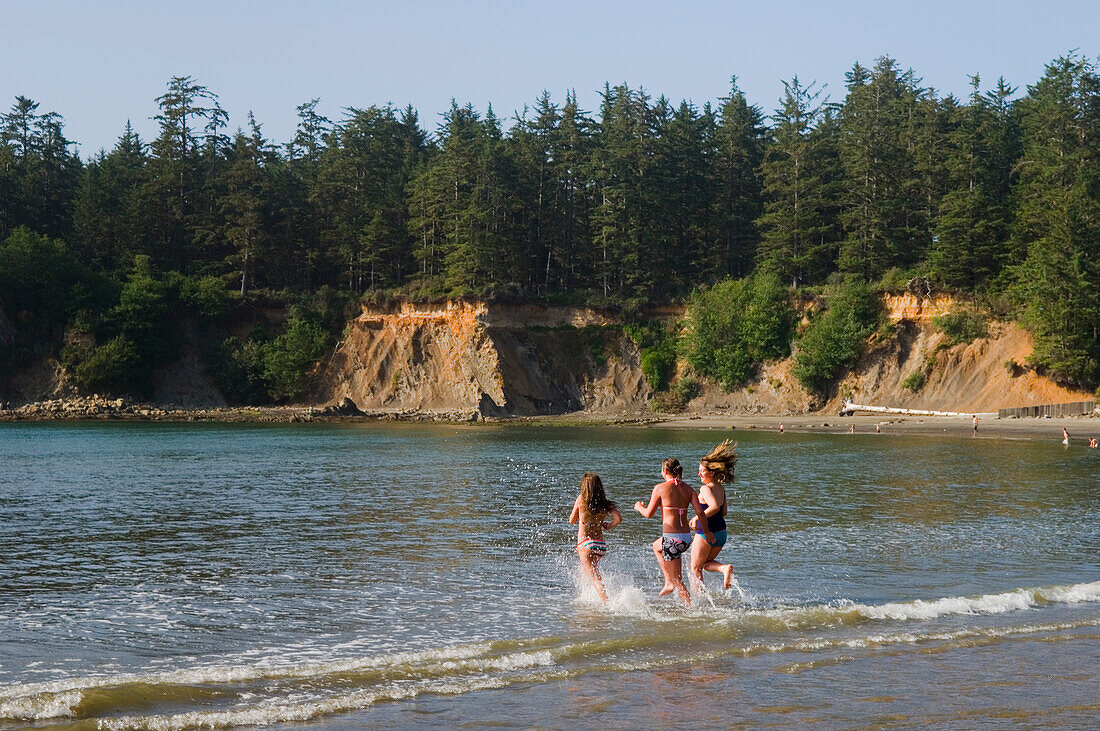 Eine Gruppe von Mädchen im Teenageralter rennt am Strand des Sunset Bay State Park an der Küste von Oregon ins Wasser.