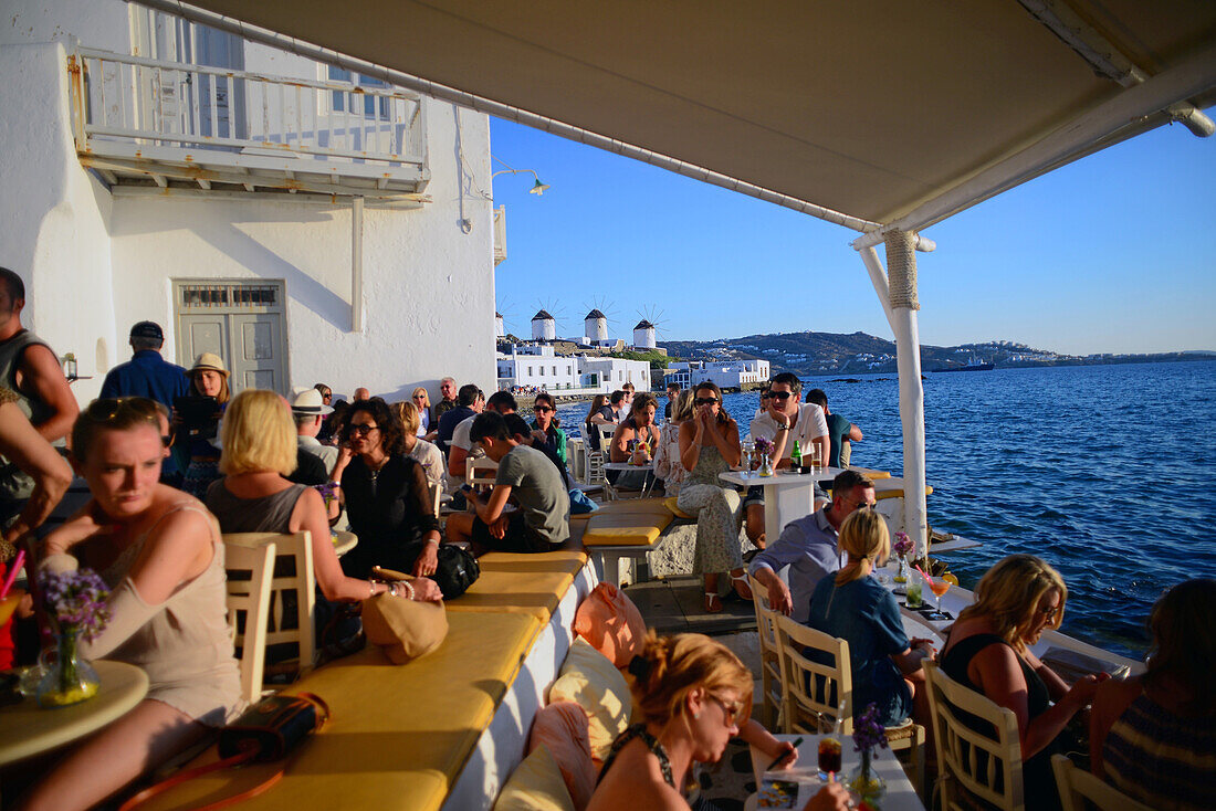 Besucher genießen den Sonnenuntergang in Mykonos von einer Terrasse mit Blick auf Windmühlen, Griechische Inseln, Griechenland