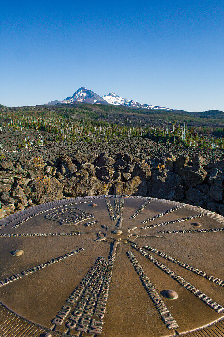 Bronze plaque pointing out mountain peaks seen from Dee Wright Observatory at McKenzie Pass in the Cascade Mountains of Oregon, with North Sister and Middle Sister volcanic peaks in the distance.