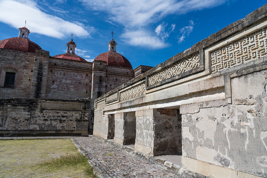 Laubsägearbeitstafeln aus Stein an den Wänden von Hof B (Viereck B) in den Ruinen der zapotekischen Stadt Mitla in der Nähe von Oaxaca, Mexiko. Dahinter befindet sich die Kirche von San Pablo. Ein UNESCO-Weltkulturerbe.