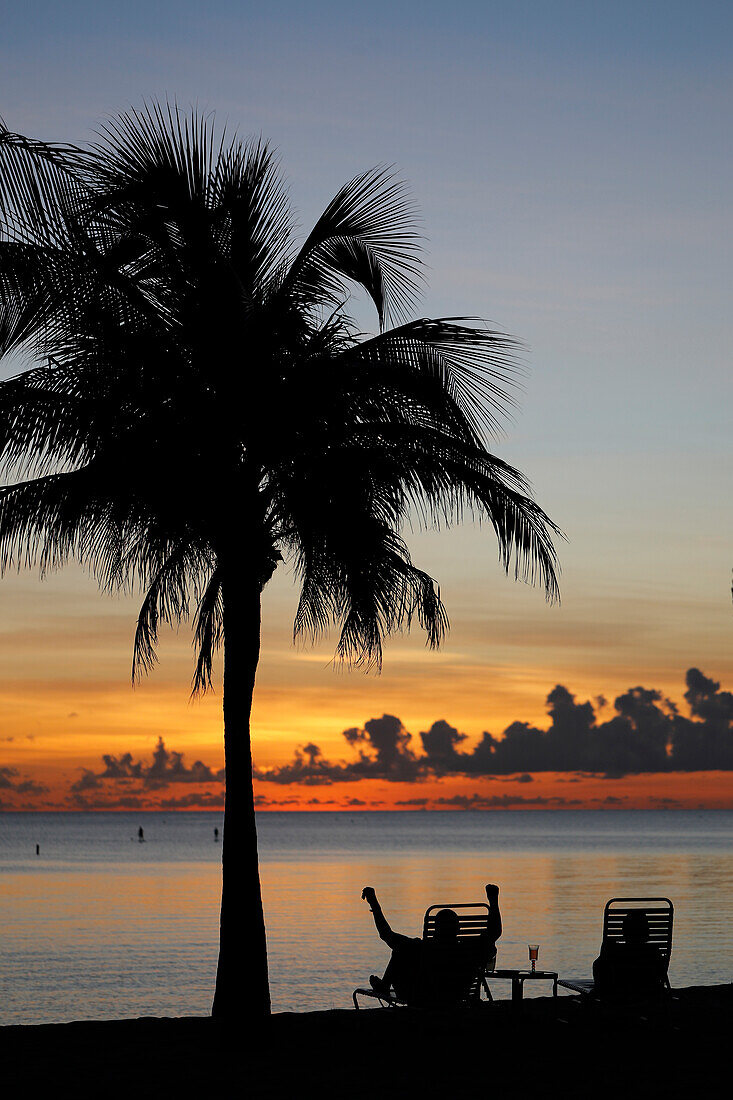 Mann im Ruhestand sitzt auf einem Stuhl bei Sonnenuntergang, tropischer Strand und paradiesische Natur, Kaimaninseln, Karibik, Mittelamerika