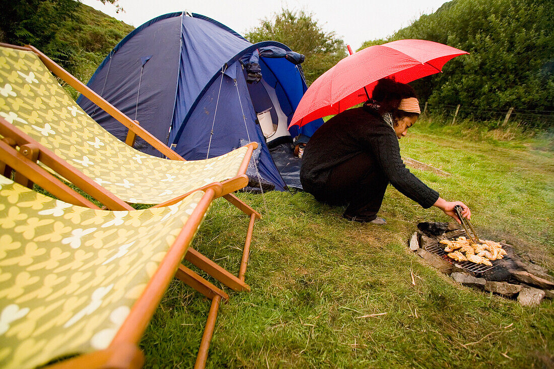 Camperin grillt Hähnchenstücke auf dem Lagerfeuer im Regen