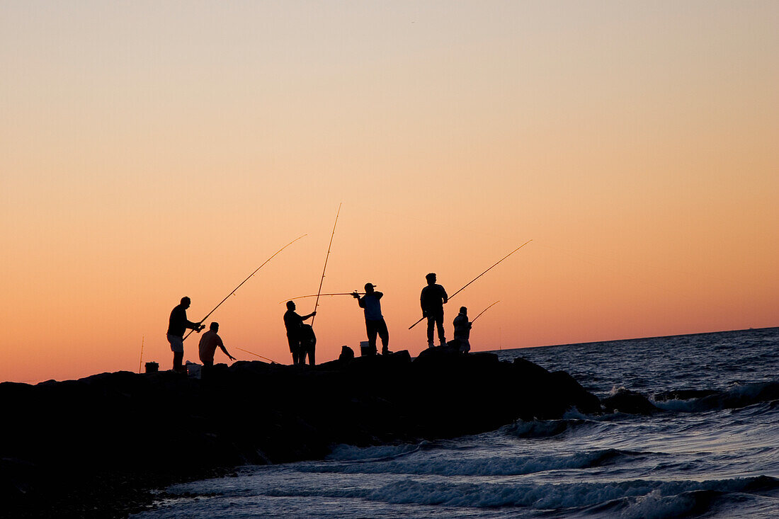 Silhouetted People Fishing Off Rocks On Beach At Sunset