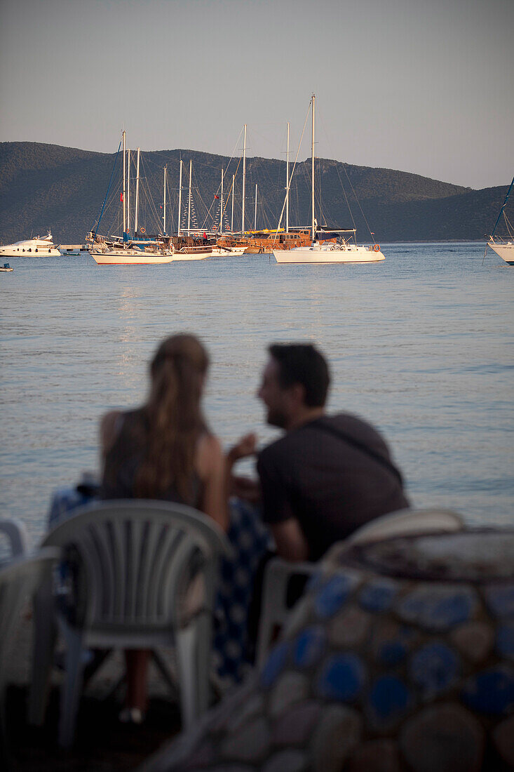 Türkei, Pärchen teilt Moment und Getränk am Strand der Ostbucht von Bodrum mit Blick auf die Gulet-Boote; Bodrum