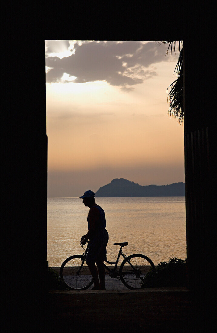 Silhouette eines Mannes, der in der Abenddämmerung ein Fahrrad entlang der Strandpromenade schiebt, Seitenansicht, Lopud, Kroatien