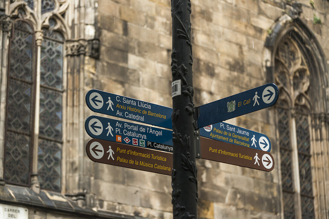 Wegweiser für Sehenswürdigkeiten an einem Pfosten mit einer alten Steinmauer und Fenstern im Hintergrund, Gotisches Viertel; Barcelona, Katalonien, Spanien.