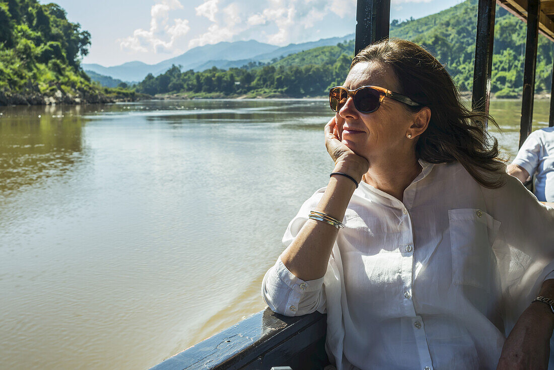 Eine Frau mit Sonnenbrille sitzt in einem Ausflugsboot und schaut aus dem Fenster, während sie den Mekong hinunterfährt; Provinz Luang Prabang, Laos