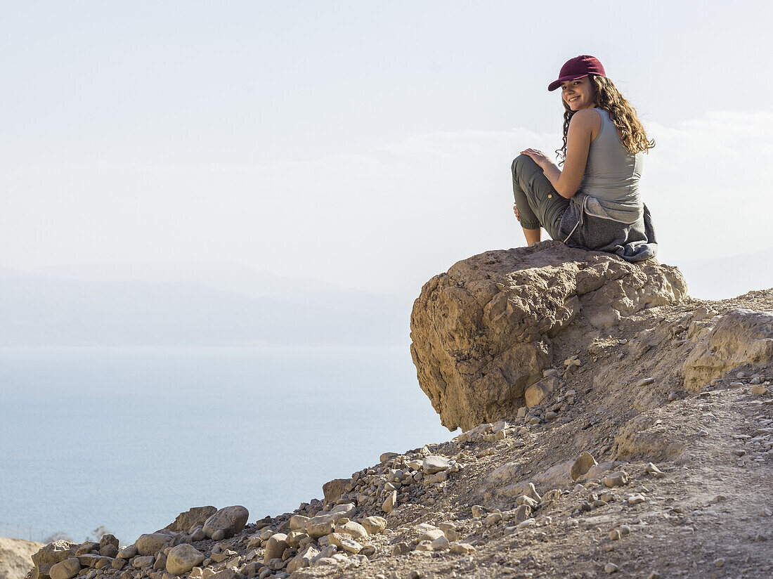 Eine junge Frau sitzt auf einem Felsen und posiert für die Kamera mit einem wolkenbedeckten Tal unter ihr, Naturreservat Ein Gedi, Distrikt Totes Meer; Region Süd, Israel