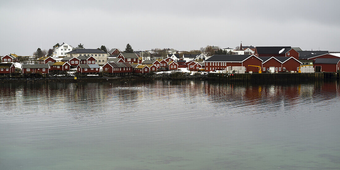 Red Buildings Along The Water's Edge Under A Cloudy Sky; Lofoten Islands, Nordland, Norway