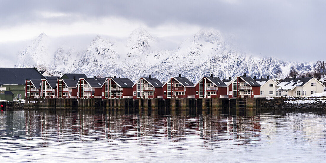 Red Buildings Along The Water's Edge With Snow Covered Mountains In The Background; Svolvar, Lofoten Islands, Nordland, Norway