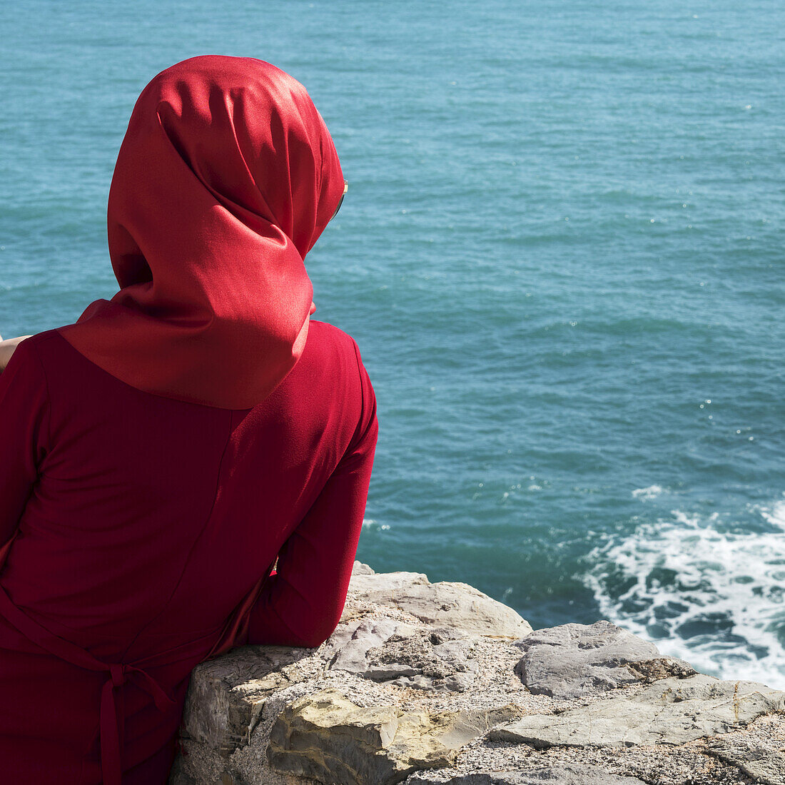 A woman wearing a red headscarf and red dress stands at a stone wall looking out at the blue water; Budva, Budva Municipality, Montenegro