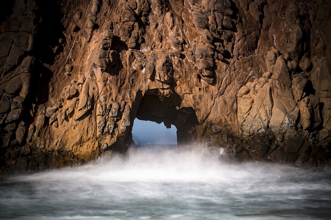 A natural arch in the rock formation along the coast with mist rising off the water; Big Sur, California, United States of America