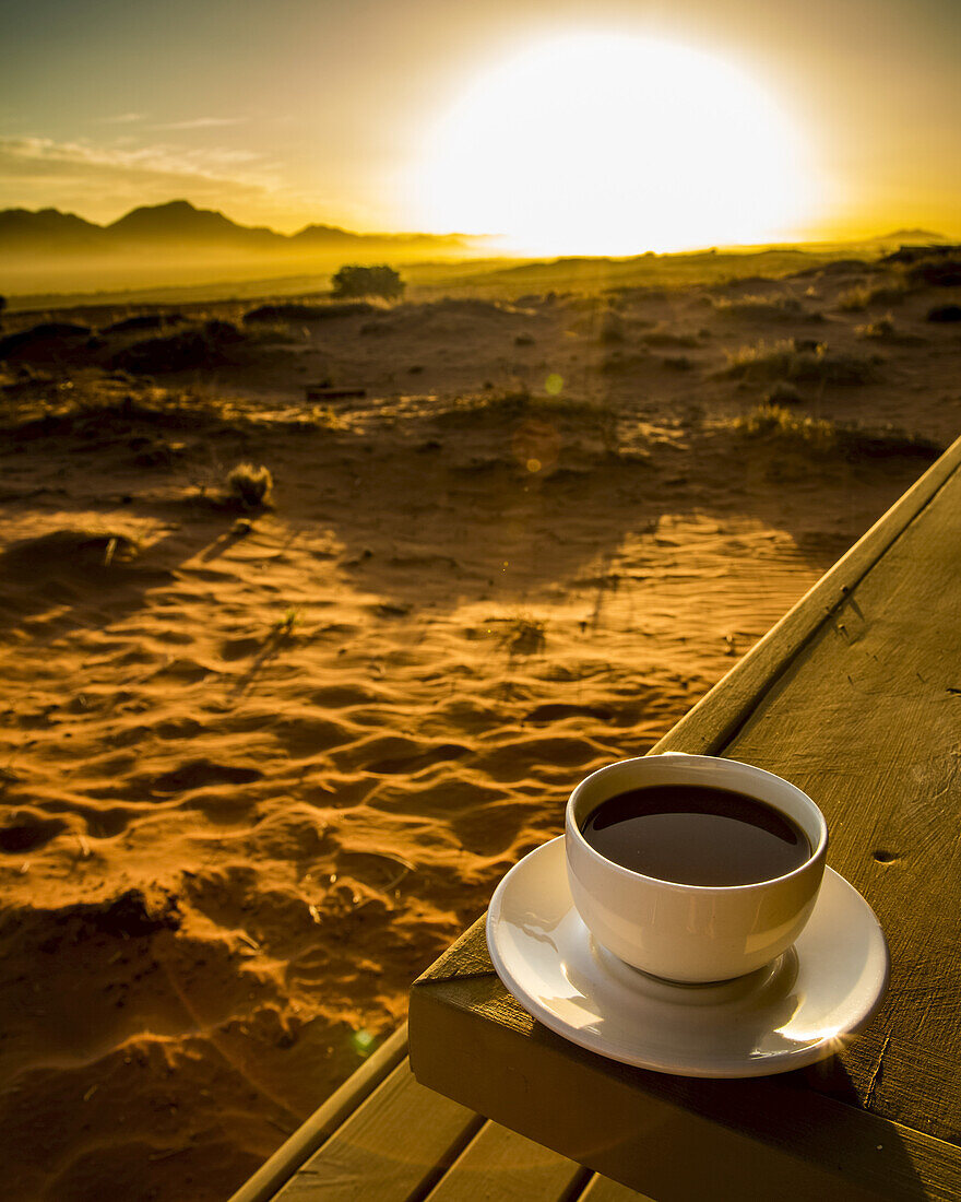 A cup of coffee sits on a wooden table with a view of the sunrise over the desert; Sossusvlei, Hardap Region, Namibia