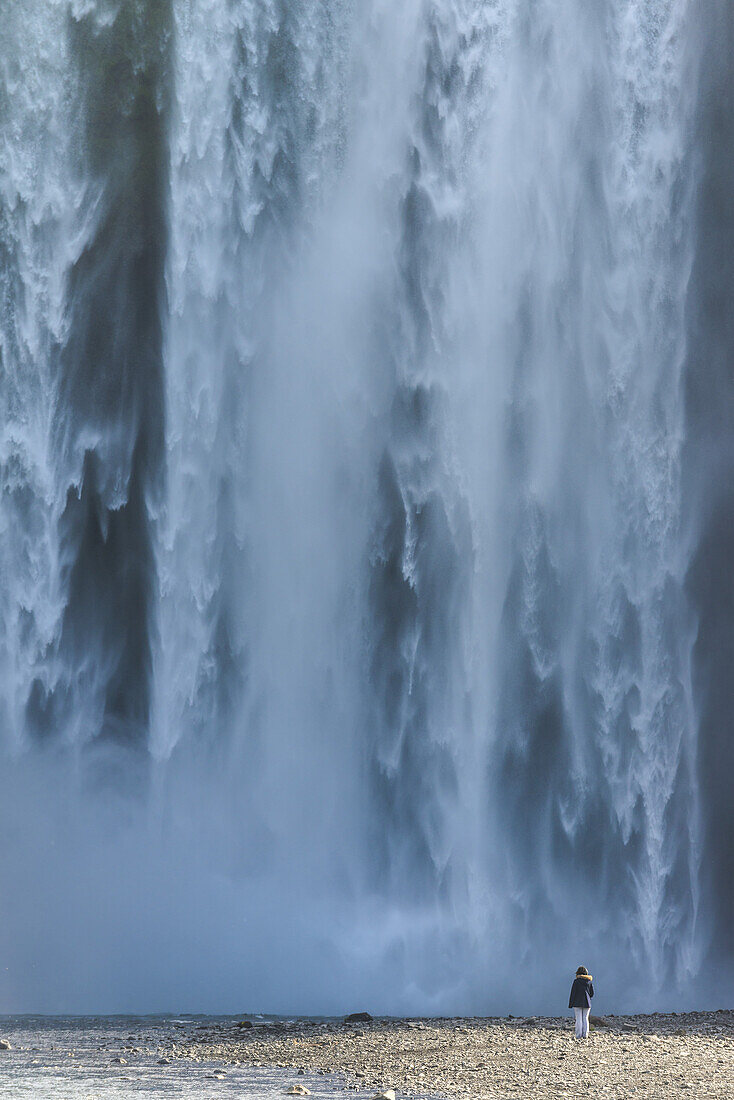 Frau steht am Fuße des Skogafoss-Wasserfalls und schaut auf das kraftvoll fließende Wasser; Island