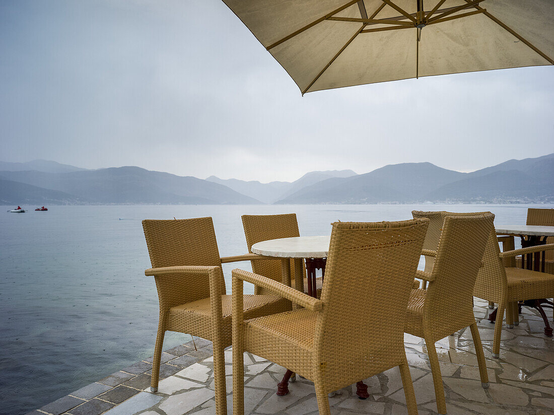 Tische und Stühle unter einem Sonnenschirm an der Uferpromenade der Bucht von Kotor; Bjelila, Opstina Tivat, Montenegro.