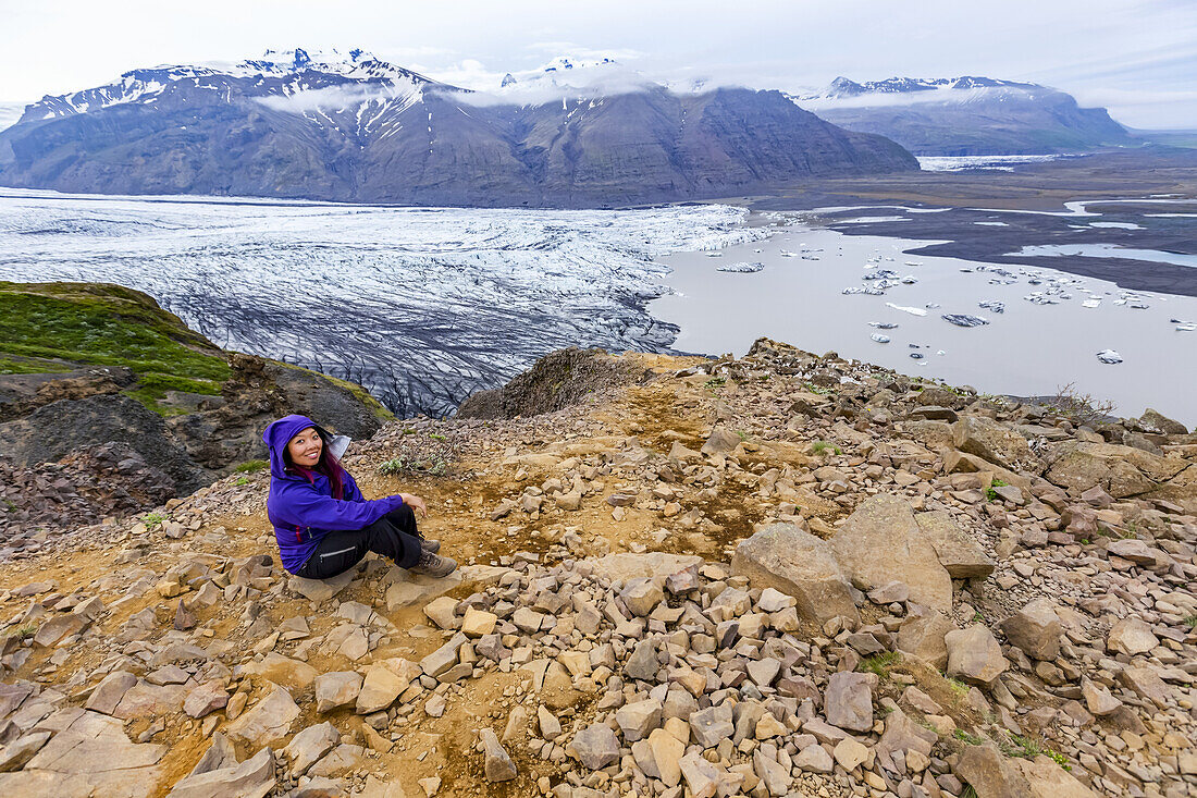 A female hiker in warm clothing poses on a mountain top overlooking the glacier lake and valley below at Vatnajokull National Park; Iceland