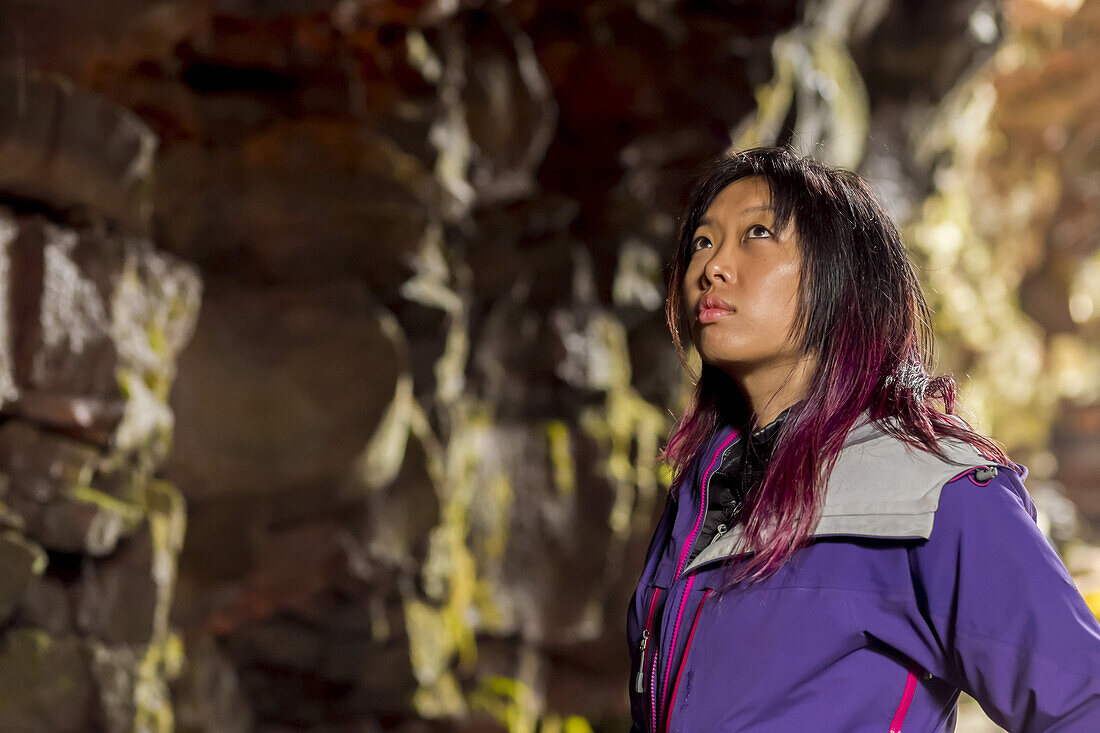 Nahaufnahme einer asiatischen Wanderin im Seitenprofil mit Blick nach oben auf die Höhlenwände in einer Lavaröhrenhöhle; Island.