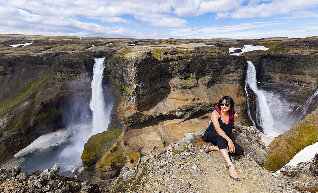 Eine junge asiatische Wanderin posiert für ein Porträt am Rande einer atemberaubenden Landschaft mit zwei Wasserfällen, die als Haifoss bekannt ist; Island