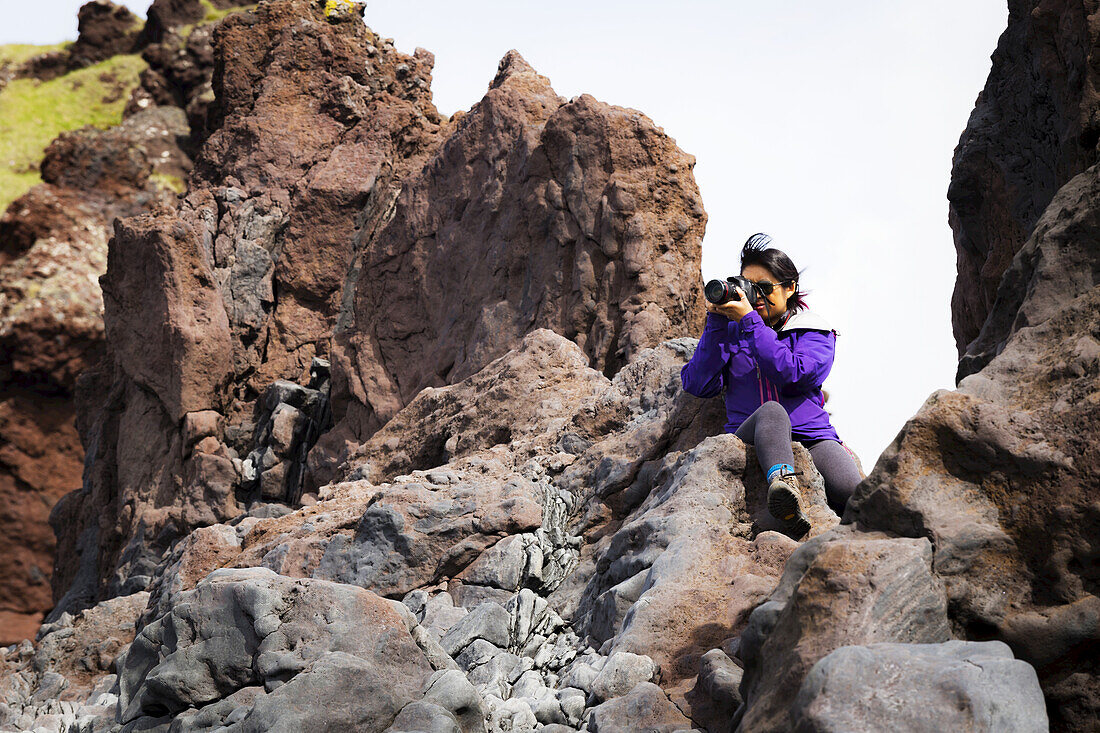 Eine Touristin sitzt auf der Spitze einer Felsformation, um ein höheres Foto von der Landschaft zu machen; Island