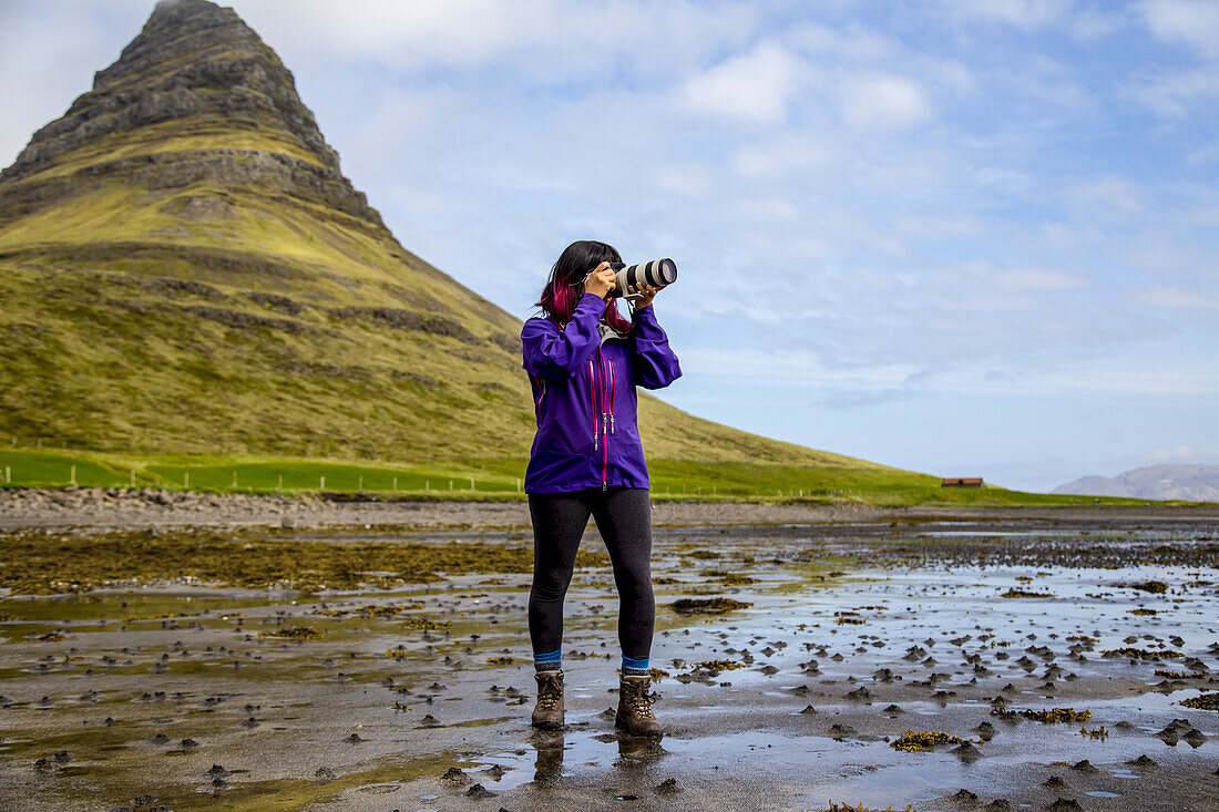 A female hiker stops on the beach at low tide to take a photo with an SLR camera in front of Kirkjufell Mountain in Snaefellsnes peninsula, Western Iceland; Iceland