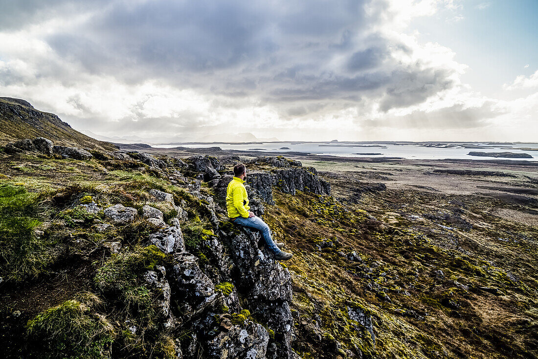 Man looks out over the ocean while sitting on a cliff; Iceland