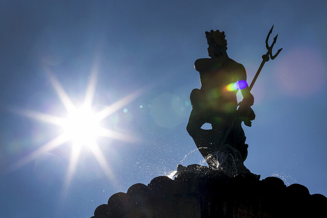 Silhouette of statue on top of fountain with sunburst and blue sky; Trento, Trento, Italy