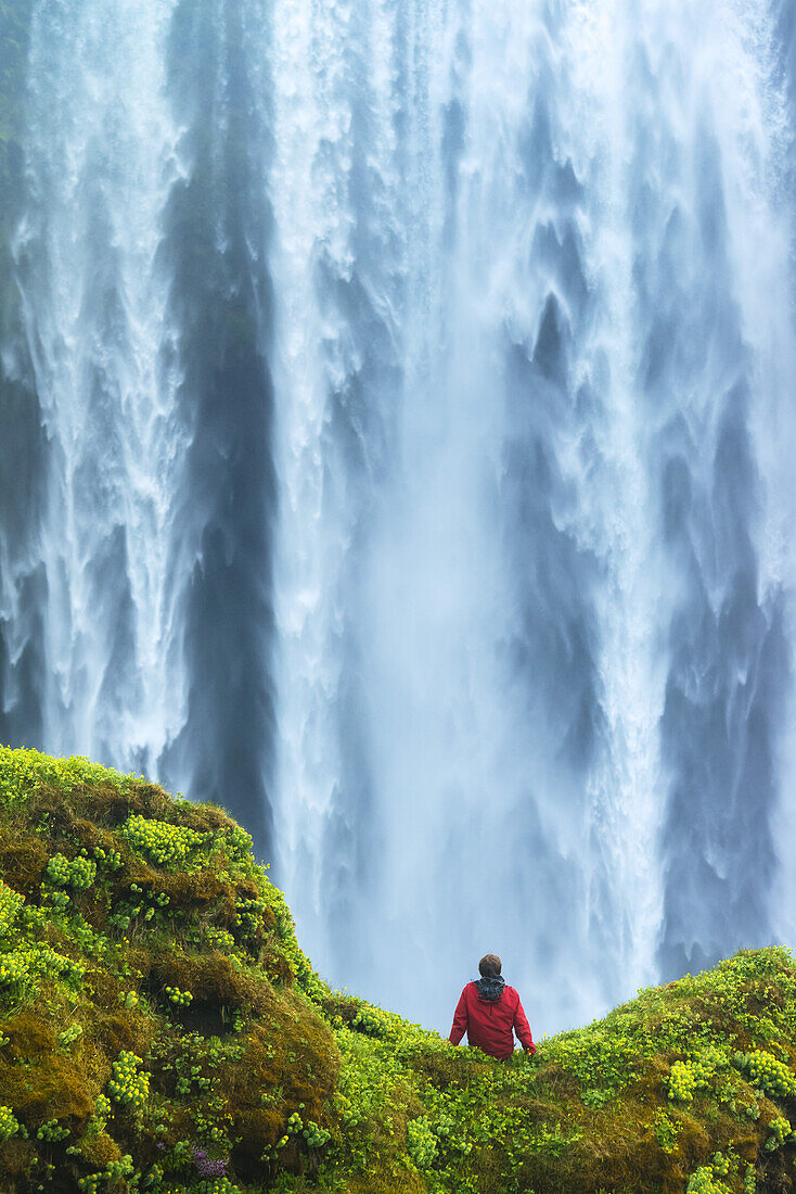 Mann sitzt auf einem moosbewachsenen Felsen am Fuße des Skogafoss-Wasserfalls; Island