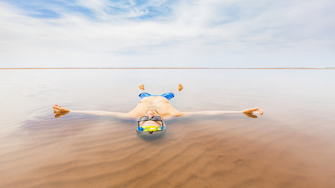 A man wearing a bathing suit floats in the shallow ocean water with his body outstretched and looking up to the sky; Prince Edward Island, Canada