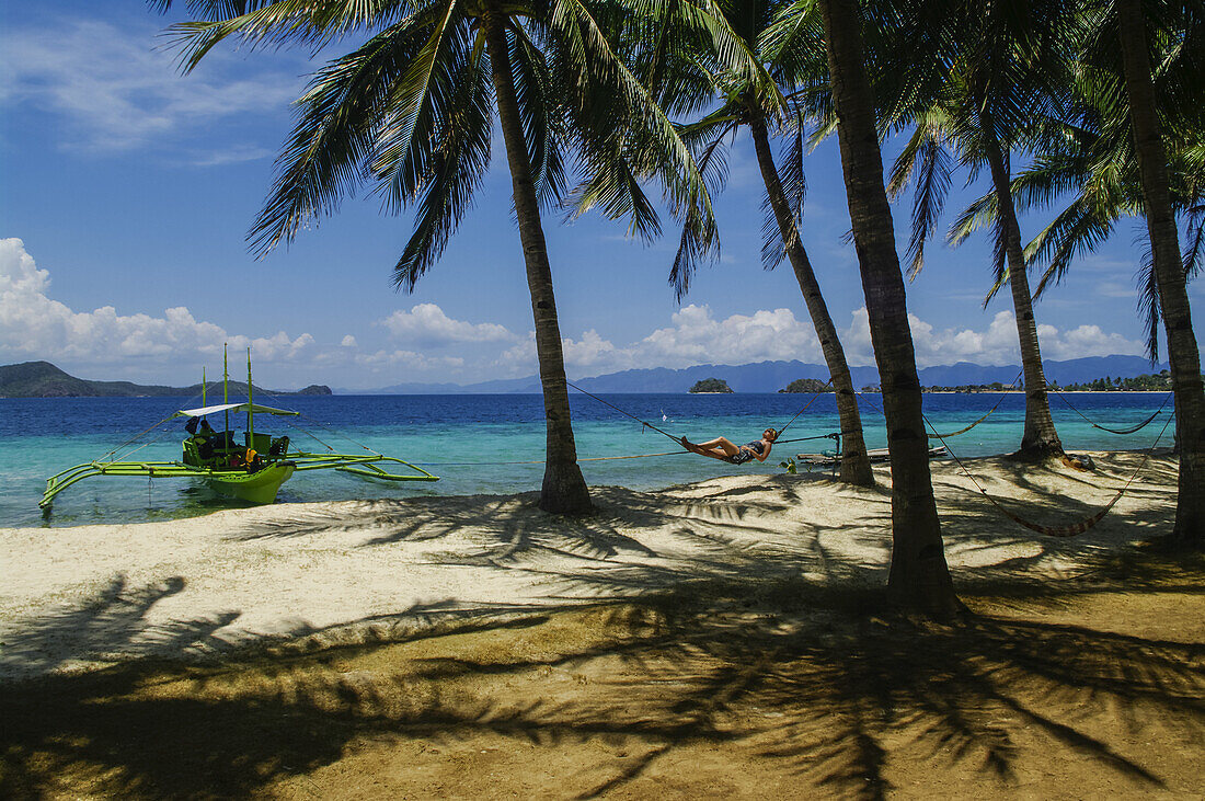 Eine Frau liegt in einer Hängematte an einem tropischen Strand mit einem Auslegerkanu, das am Ufer vertäut ist; Andamanen, Indien.