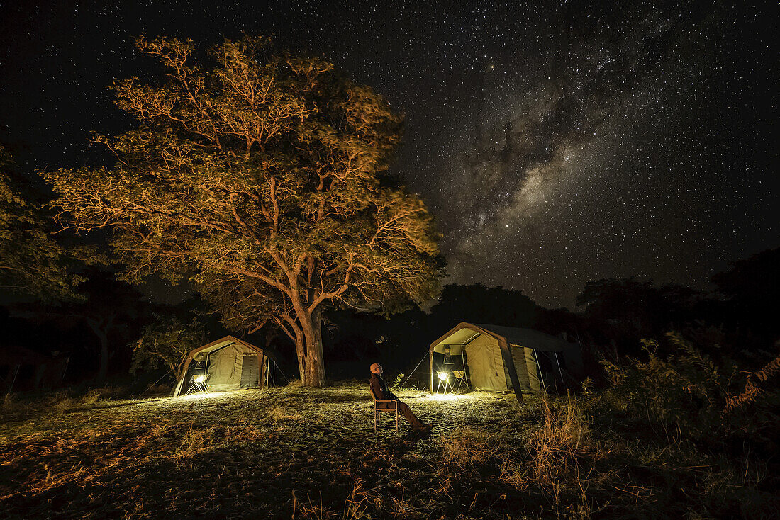 Die Milchstraße am Himmel, darunter ein Zelt in einem Buschcamp, während ein Mann sitzt und in den Himmel schaut; Botswana