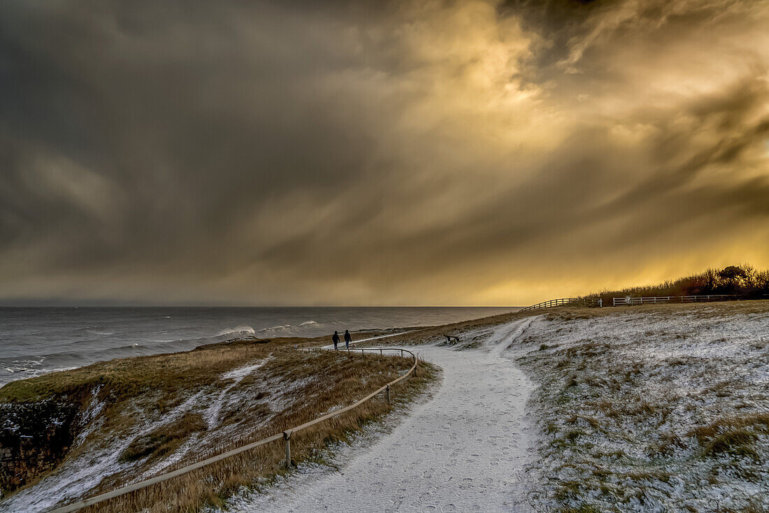 Two figures walking on the snowy path along the coast at dusk; South Shields, Tyne and Wear, England